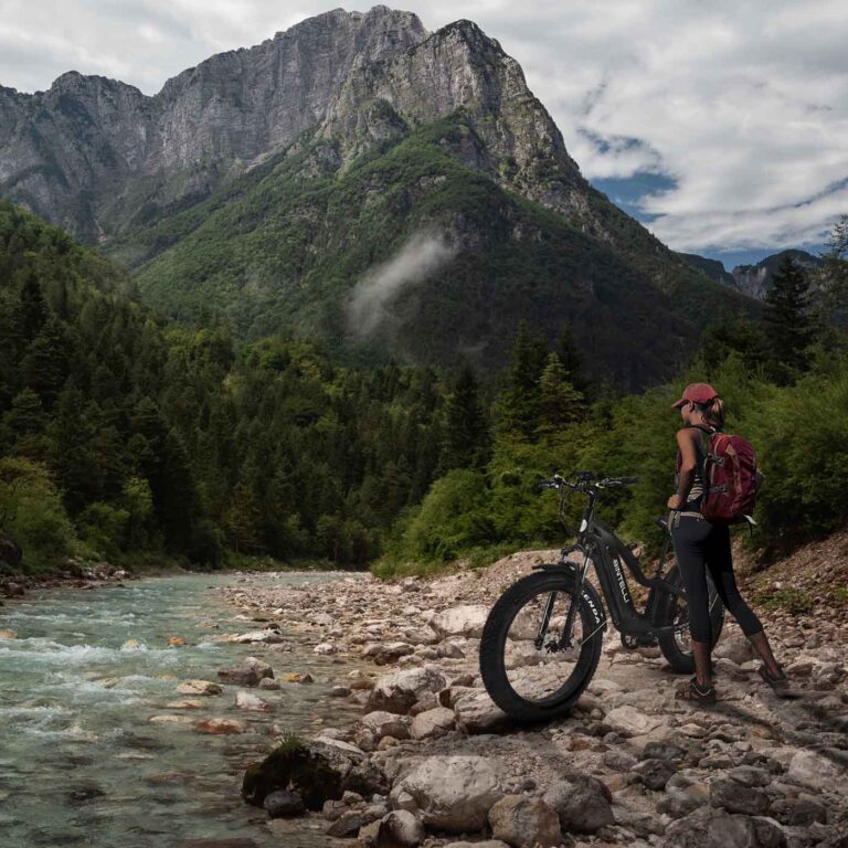 A woman standing next to the bintelli electric black black bike near a river.